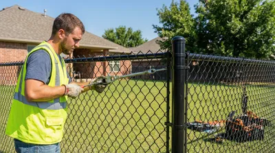 Professional fence installer in safety vest using tension tools to secure black chain-link fencing to a post in a residential backyard with brick homes visible in background.