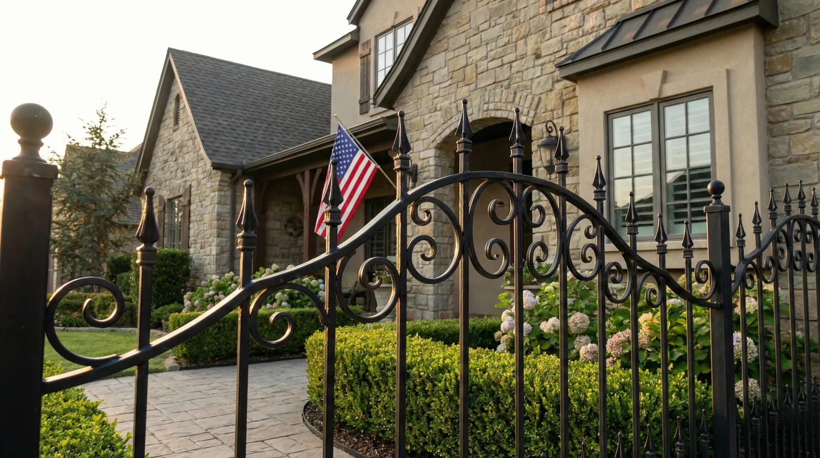 Ornate black iron fence with decorative scrollwork stands in front of a luxury stone home with American flags displayed, featuring manicured landscaping and a paver walkway.