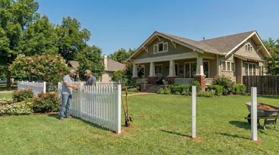White picket fence installation in progress outside a craftsman-style bungalow with tan siding and brick accents. Fresh fence posts are set in front of a well-maintained lawn with flowering shrubs and trees.