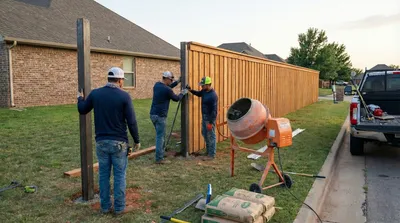 Construction crew installing tall wooden privacy fence panels alongside a brick residential home, with a cement mixer and building materials visible on the ground
