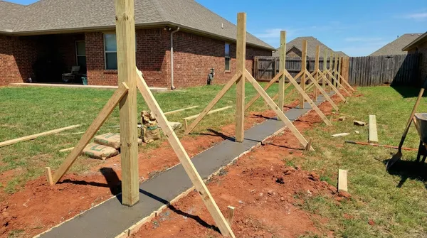 Wooden fence posts temporarily braced with diagonal supports stand in freshly poured concrete along a residential backyard with red clay soil and brick homes visible in the background.