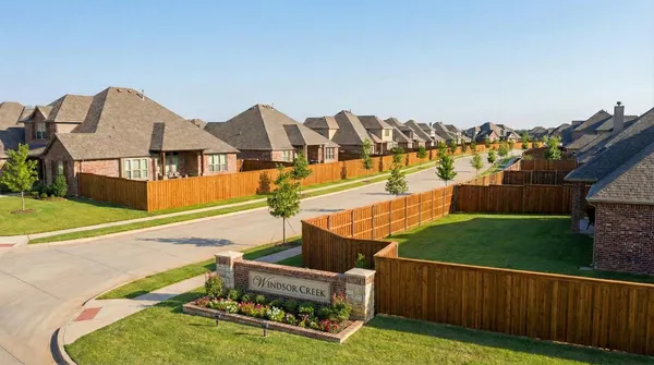 Suburban neighborhood entrance marked by Windsor Creek sign, featuring brick homes with uniform wooden privacy fences, manicured lawns, and young trees lining concrete sidewalks under a clear blue sky.