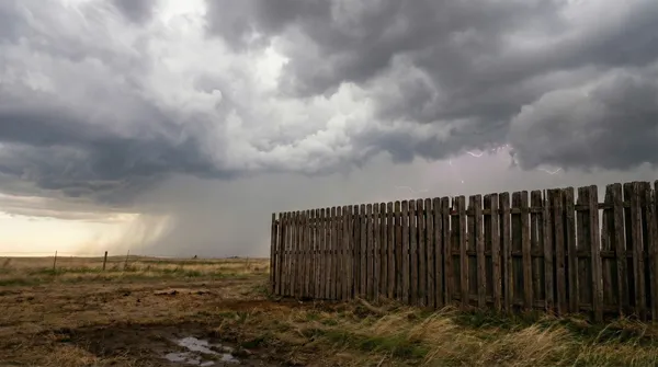 Lightning strikes illuminate dark storm clouds above a weathered wooden privacy fence stretching across an open field in Tulsa, with rain visible in the distance and wind-blown grass in the foreground.
